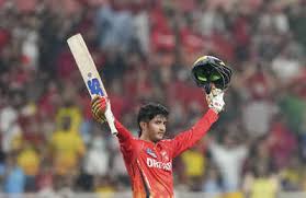 A cricketer in a red jersey and helmet raises his bat in celebration, surrounded by a cheering crowd.