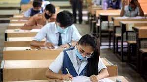 Students sitting in an examination hall, focused on writing answers in their answer sheets while wearing masks as a safety measure.